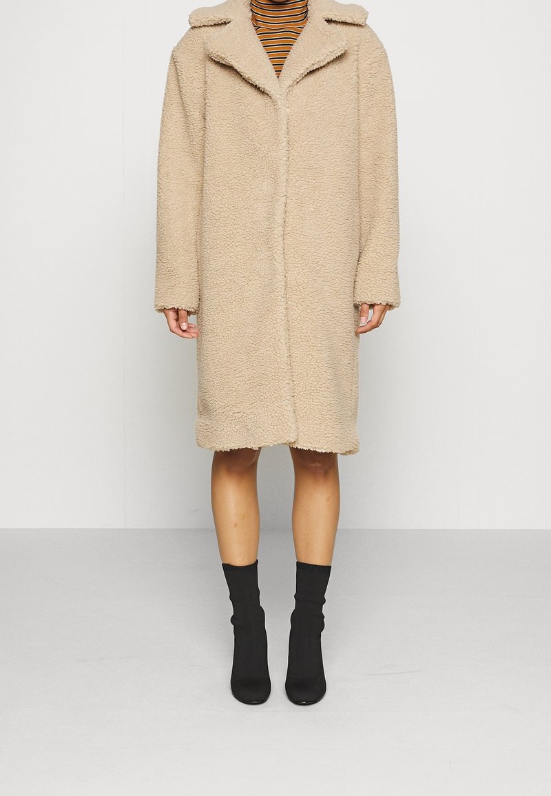 Beiger Kunstpelzmantel mit Kragen, langen Ärmeln und geradem Schnitt, der bis zur Mitte der Oberschenkel reicht. Getragen über einem gestreiften Oberteil und schwarzen Socken.