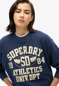 Sweatshirt marine avec logo brodé blanc et or "SUPERDRY" et "ATHLETICS". Col rond classique avec poignets côtelés. Texture douce.