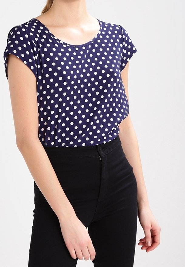 Femme portant un chemisier bleu marine à manches courtes avec des pois blancs, rentré dans un pantalon noir taille haute.