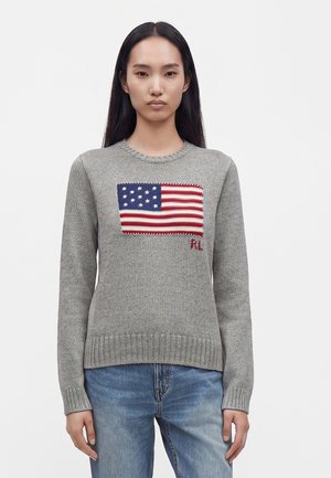 En person med langt sort hår iført en grå strikket sweater med et påsyet amerikansk flag og blå jeans, stående mod en ensartet baggrund.