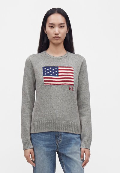 Une personne aux longs cheveux noirs porte un pull en tricot gris avec un drapeau américain cousu et un jean bleu, debout devant un fond uni.