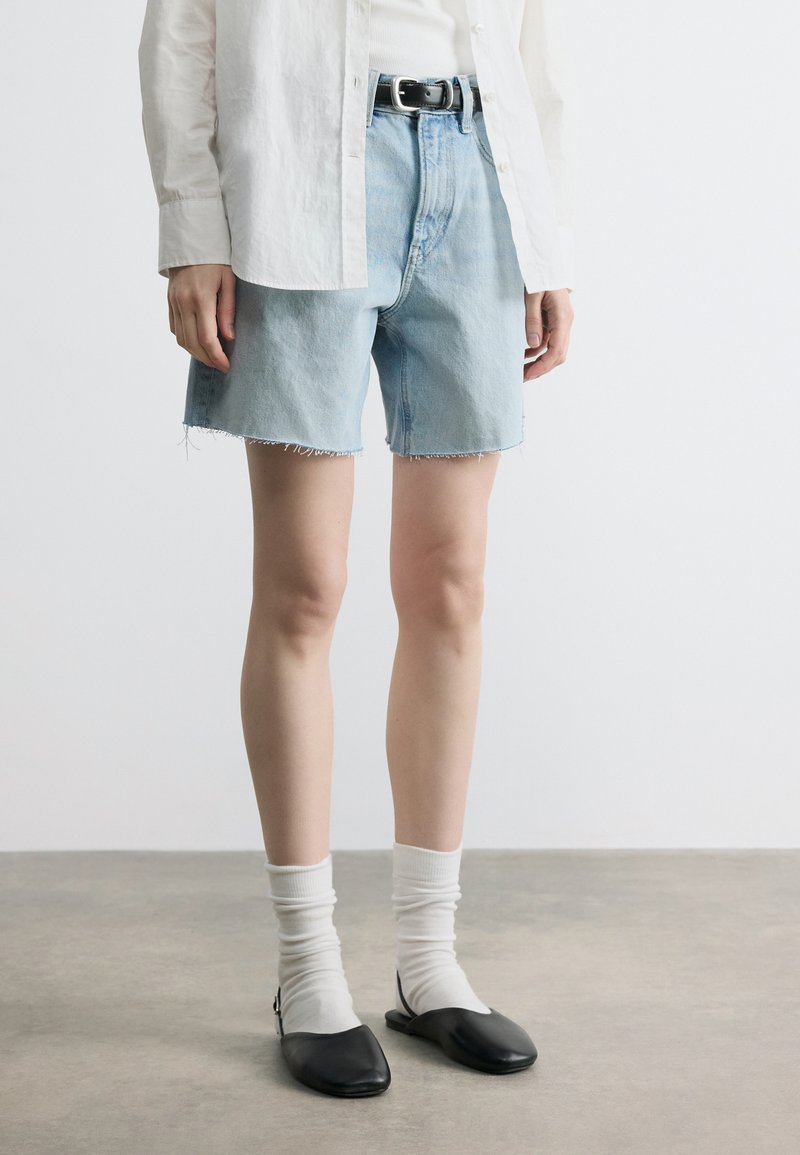 Άτομο που φοράει ανοιχτό μπλε τζιν σορτς, λευκό πουκάμισο με μακριά μανίκια, λευκές κάλτσες και μαύρα επίπεδα παπούτσια, στέκεται σε γκρίζο πάτωμα.