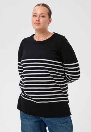 Femme aux cheveux attachés porte un pull noir à rayures horizontales blanches et un jean bleu, debout devant un fond clair uni.