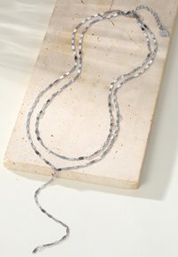 Collier argenté à plusieurs couches avec des maillons allongés et texturés. Il comporte une chaîne délicate avec un fermoir et un rallonge ajustable. Placé sur une surface claire.