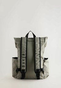 Sac à dos en tissu gris avec des bretelles noires, design à rabat, poches latérales et détails de logo. Matériau durable avec une texture lisse.