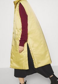 Gul satinväst i lång modell med hög krage och sidofickor, lagd över en vinröd långärmad skjorta, tillsammans med en mörk kjol och ankelstövlar.