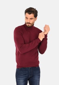 Maglione a collo alto bordeaux con polsini e orlo a costine, design aderente, indossato con jeans blu, che mette in mostra una trama a maglia liscia.
