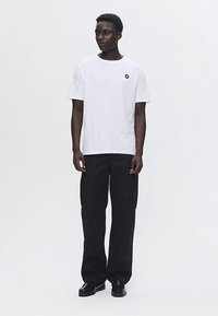 Mladenič stoji, oblečen v belo majico z majhnim črnim logotipom in ohlapnimi črnimi hlačami, pred enostavno belo ozadje.