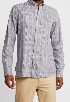 Mann trägt ein schwarz-weiß kariertes Hemd und beige Hose, steht vor einem einfarbigen hellen Hintergrund.