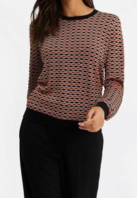 Neulottu pitkähihainen pusero, jossa aaltoileva kuvio punaisessa, oranssissa ja mustassa kermaisella pohjalla sekä mustat resorikaulukset ja hihansuut.