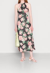 Robe maxi florale avec un fond noir, ornée de fleurs roses et vertes. Design à col halter, assortie à un sac vert clair et des sandales blanches.