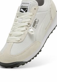 Baskets avec une tige en tissu blanc et des accents en daim beige, ornées d'un pendentif perle brillant, de lacets argentés et d'une semelle en caoutchouc noire.