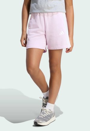 Persoon draagt lichtroze Adidas korte broek, grijs-witte sneakers, witte sokken, staand op een lichte achtergrond.