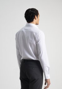 Camisa blanca con finas rayas verticales, de manga larga y cuello puntiagudo, combinada con pantalones oscuros. Textura suave, diseño clásico.