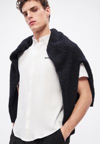 Balti marškiniai su mažyte juoda „BOSS“ logotipu, apnuoginti po puriu juodu drabužiu. Paprastas dizainas ir klasikinis apykaklė.