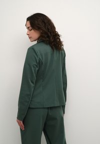 Groene sportjack met een getailleerd ontwerp, zichtbare naden en lange mouwen. Zachte textuur met een kraag en een iets langere achterkant.