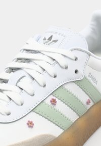 Baltas ādas sporta kurpes ar zaļām un baltām svītrām, ziedu rotājumi sānos un gumijas zole. Redzams Adidas Samba zīmols.