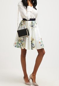 Jupe A-line fleurie en tissu blanc, ornée de fleurs multicolores, associée à un sac crossbody noir et des escarpins neutres.