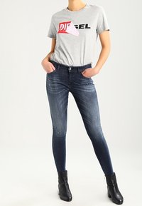 Camiseta gris con un logo rojo y blanco, combinada con jeans ajustados de color azul oscuro y botas negras hasta el tobillo. Conjunto informal sin patrones visibles.