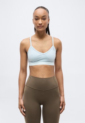 Žena s tmavými vlasmi zviazanými do copu, oblečená v svetlomodrom športovom podprsenke a hnedých legínach s vysokým pásom, stojaca na čistom pozadí.