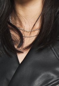 Indossate collane argentate a più livelli, con una catena spessa e una catena sottile, contro un top in pelle nera. I capelli coprono parzialmente i gioielli.