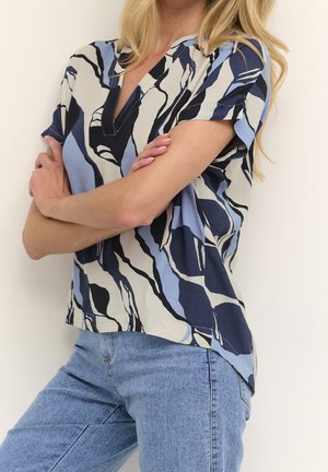 Femme aux bras croisés portant un chemisier à manches courtes à motif abstrait bleu, noir et blanc, et un jean bleu clair, devant un fond uni.