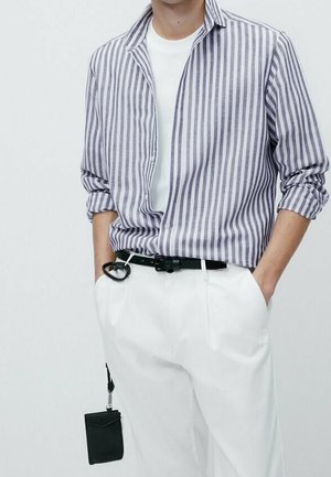 Homme portant une chemise rayée gris et blanc sur un t-shirt blanc, pantalon blanc, ceinture noire, mains dans les poches et un portefeuille noir suspendu.