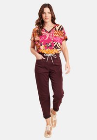 Donna con una blusa a fantasia multicolore vivace, pantaloni bordeaux con lacci e sneakers metallizzate in oro rosa che cammina in avanti.