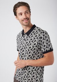 Kortärmad polo med ett svartvitt mönster av löv, med svart krage och kontrasterande ärmslut. Tillverkad av ett mjukt tyg.