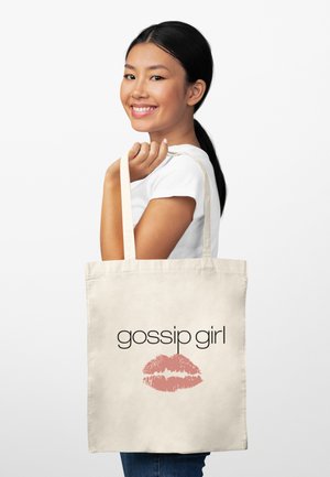 Uśmiechnięta kobieta o czarnych włosach związanych w koński ogon trzyma beżową torbę z napisem „gossip girl” oraz różowym odciskiem szminki na białym tle.