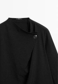 Giacca in misto lana nera con colletto alto, chiusura asimmetrica e dettaglio a un bottone. Tessuto morbido con design minimalista.