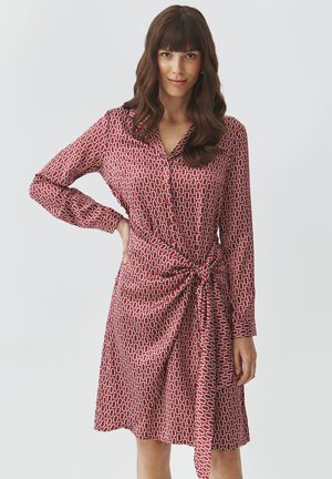 Robe rouge avec un design à motifs comportant des ovales entrelacés. Les manches longues et le détail de taille nouée créent une silhouette structurée. Texture de tissu lisse.