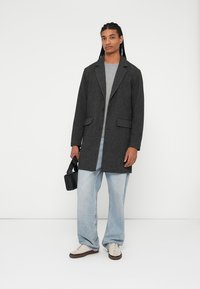 Grijs wollen blend jasje met notch-lapels, enkele knoopsluiting en twee voorkruistassen. In combinatie met een grijze trui en lichtblauwe spijkerbroek.