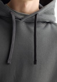 Sudadera gris oscuro con una textura suave, que cuenta con un bolsillo canguro en la parte delantera y cordones en un gris más oscuro. Diseño simple y minimalista.