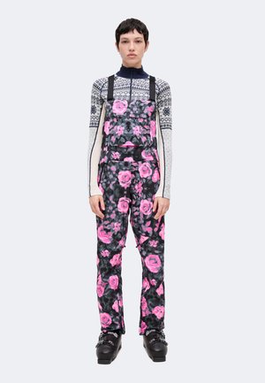Personne debout portant des chaussures de ski noires, une combinaison fleurie noire et rose, et un haut à manches longues à motifs géométriques bleus et blancs.