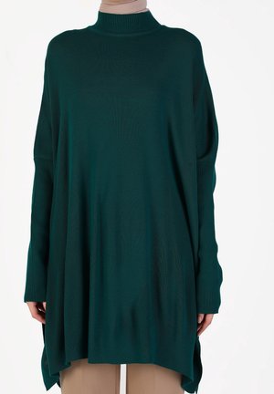 Pull à col roulé en maille côtelée vert foncé avec de longues manches tombantes et des fentes latérales, porté sur un pantalon beige.