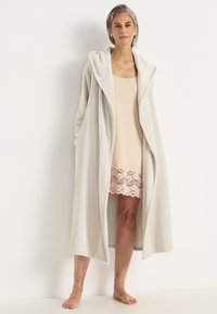 Ljusgrå, huva badrock i mjukt, plyschmaterial; långa ärmar, öppen front och sidofickor, bärs över en beige klänning med spetsdetaljer.