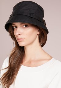 Chapeau seau noir en tissu résistant à l'eau, avec un large bord, une sangle en cuir ajustable avec des éléments en or, et une finition lisse.