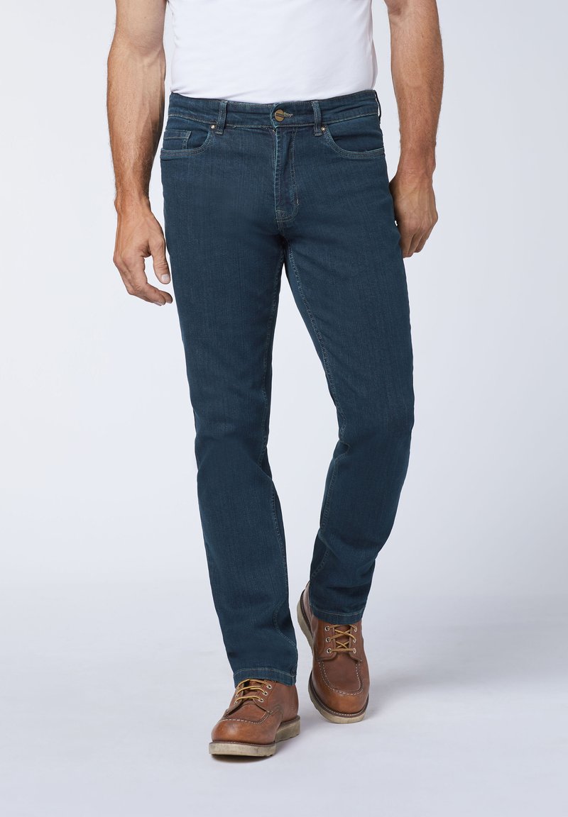Dark blue denim jeans with a straight fit, featuring a button closure, zipper fly, and five pockets. Worn with brown lace-up shoes.