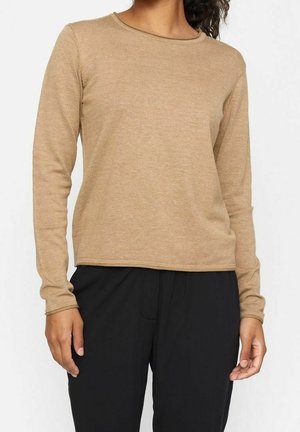 Femme portant un haut en tricot à manches longues couleur camel associé à un pantalon noir ample, debout avec les bras détendus le long du corps.