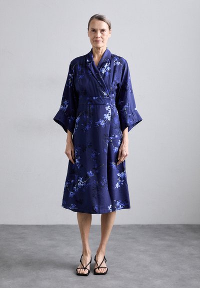Marine blomster kimono kjole med brede ærmer, vridningsdesign og blå blomstermønstre. Blød tekstur, længde til midten af læggen, parret med sorte sandaler.