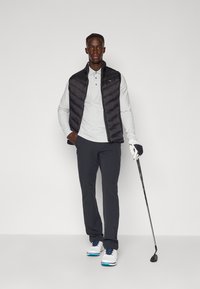 Colete acolchoado preto sobre uma polo cinza, calças escuras e sapatos de golfe brancos com detalhes azuis. Segurando um taco de golfe, usando uma luva branca.