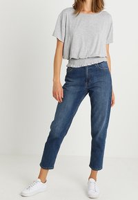 Šedý krátký rukávový cropped top se shromážděním na spodním lemu spojený s vysokým pasem modrých džínsů a bílými teniskami. Hladká tkanina, uvolněný střih.