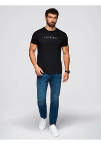 Sort bomulds T-shirt med farverig broderet tekst "amure" på brystet, parret med blå jeans og hvide sneakers.
