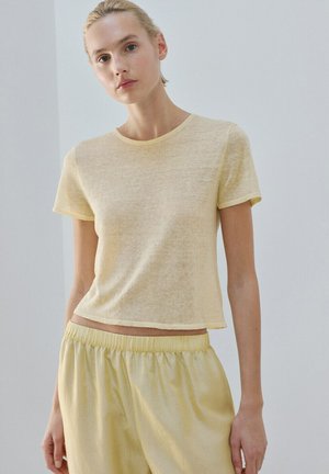 Donna che indossa una maglia trasparente giallo pallido a maniche corte e pantaloni larghi abbinati, in piedi davanti a uno sfondo chiaro e uniforme.