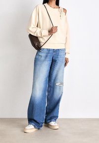 Sudadera beige con diseño asimétrico y lazo lateral, combinada con vaqueros azules desgastados de pierna ancha y zapatillas blancas. Bolso bandolera marrón.