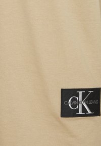 Béžová tkanina s hladkým povrchem, obsahující černou obdélníkovou etiketou s bílým textem "CALVIN KLEIN JEANS" a stylizovaným logem "CK".