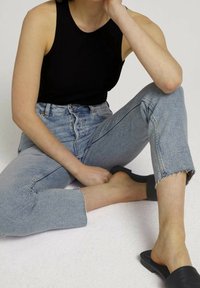 Svart ribbad linne, högmidjade ljusblå jeans med fransade fållar, och svarta tofflor. Sittande pose mot en ljus bakgrund.