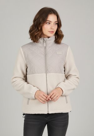 Woman wearing beige fleece jacket with quilted upper section and zip closure, standing against plain gray background, hands clasped in front.