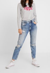 Szara koszula z długim rękawem z czerwonym logiem "Tommy Hilfiger", w zestawie z jasnoniebieskimi dżinsami z przetarciami, posiadającymi dziury i prostą nogawką. Białe trampki.